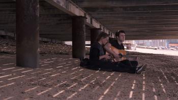 Niall McNamee sits next to Shana Swash as he plays guitar under a bridge