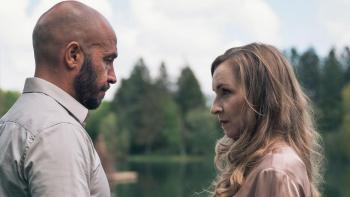 A man and woman in profile, talking against a forested background.