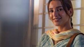 Maa Behen: Smiling Indian woman in traditional attire, standing near a window.