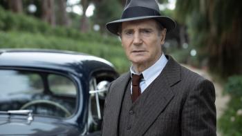 Marlowe Movie: Philip Marlowe (Liam Neeson), in 1930s period clothing and a fedora, stands next to a car and looks pensive