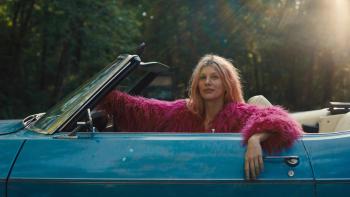 Marmalade: Camilla Morrone wears a bright pink jacket and sits in the driver's seat with her arm over the side of a blue convertible with its top down