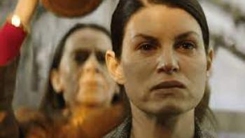 Close-up of a woman looking tense as another woman behind her raises an unknown object overhead.