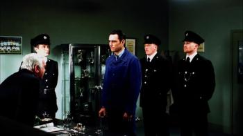 Mean Machine: A man in a blue prison uniform stands before a seated official, flanked by three guards in dark uniforms and caps