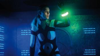 Meander: A young White blond woman holds up an illuminated bracelet in a dark tunnel, bathed in blue light.