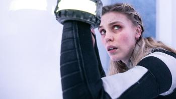 Meander: A young White blond woman holds up an illuminated bracelet, crouched in a small, bright white space.