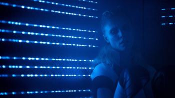 Meander: A young White blond woman leans back against a wall, with blue light shining through small slats in the darkness.