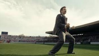 Mexico 86 Movie: Diego Luna in suit energetically cheering on a soccer field with a crowded stadium.