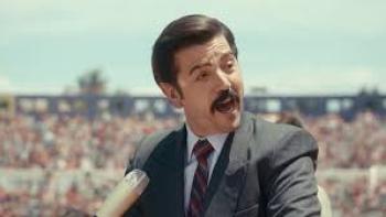 Mexico 86 Movie: Diego Luna in a suit speaking into a microphone at an outdoor event.