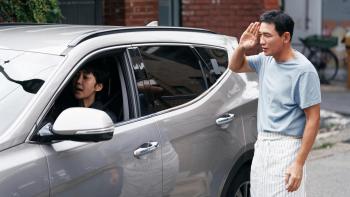 Mission: Cross movie: Korean woman drives off in minivan left, while Korean man in apron right salutes her