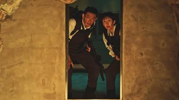 Mission: Cross movie: Korean man, woman, both with guns and in riot gear, center, peer through darkly lit doorway