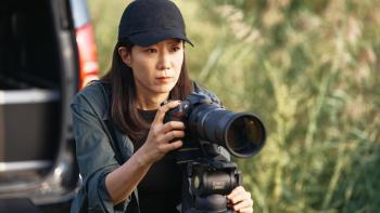Mission: Cross movie: Korean woman in black baseball cap left center holds large camera steady on a tripod, grass behind her