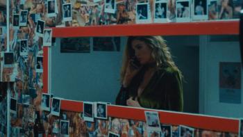 Monica Movie: A woman on the phone through the reflection in a rectangular mirror hanging on a wall full of Polaroid pictures