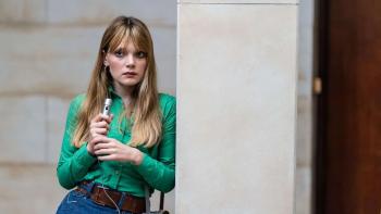 Mr. Car and the Knights Templar movie: A female reporter holding a microphone leans against a wall while wearing a green shirt. She has a serious look on her face