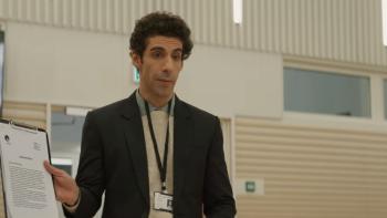 Mrs. Chatterjee vs. Norway: A young Indian man stands in beige room wearing a dark jacket holding a clipboard