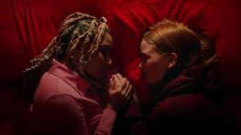 My Animal Movie: Two young women, one Black and one White, lie on a bed with red sheets, holding hands while facing each other