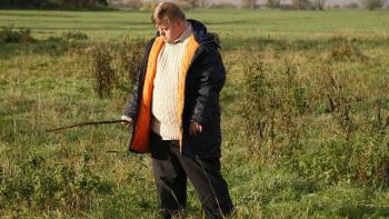 My Feral Heart: Steven Brandon wears a thick winter coat and stands in a field holding a stick and looking down at the ground