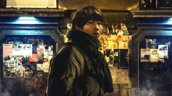 My Name is Loh Kiwan movie: Korean man in winter coat and scarf center walks down street with public poster boards behind him