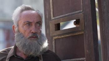 My Neighbor, Adolf: A White bearded-man stands next to a wooden door, his eyes in line with a peephole