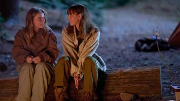 My Old Ass Movie: Young Elliott and older Elliott sit on a log by a campfire, smiling at each other