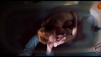 My Animal Movie: A young redheaded woman clutches her knees tightly in a bathtub while a woman's hands are seen, washing her cuts and bruises