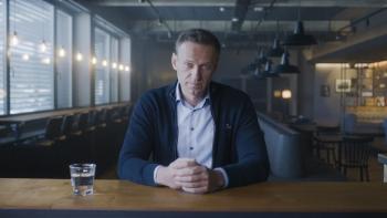 Navalny: A Russian white man center with hands clasped sits facing camera with glass of water on left