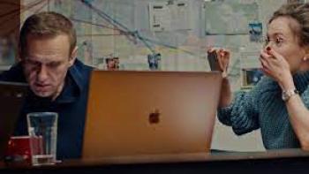 Navalny: A Russian white man sitting on left looks down while a Russian white woman on right with hand over her mouth films him with camera phone, while an Apple laptop sits between them