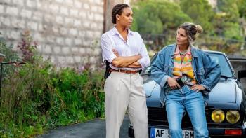 Nice Girls movie: Black French woman in white center stands talking to White French woman in jeans sitting on car hood