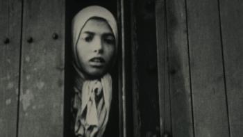 Night and Fog: A woman in a head scarf looks through an opening in a wooden door