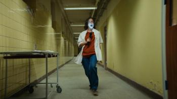 Nightwatch: Demons Are Forever movie: Danish white woman in white lab coat, headphones, and flashlight sings in hallway