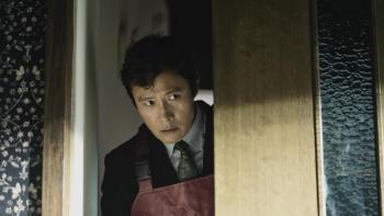 No Other Choice Movie: Wearing burgundy-colored waders, Man-su peeks through a doorway, looking wary
