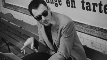 Nouvelle Vague Movie: In dark glasses, Godard sits on a subway bench, smoking and working on pages for his movie