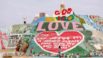 Off the Record movie: Ryan Hansen stands atop an art display on a hill sayihg "God Is Love"; Rainey Qualley looks up at him