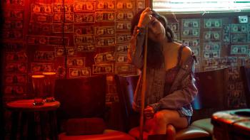 Off the Record movie: Rainey Qualley sits in front of a dollar-bill-decorated wall, holding a pool cue, with a jean jacket falling off her shoulder