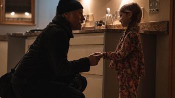 Old Guy movie: Christoph Waltz, dressed in black (including a beanie), squats down to talk to a little girl