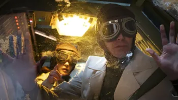 Our Times movie: Mexican woman background, man foreground, both wearing aeronautical headgear in time capsule machine, yellow light above them