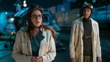 Our Times movie: Mexican woman left, man right, both in lab coats and aeronautical headgear stand in front of large time capsule machine at night