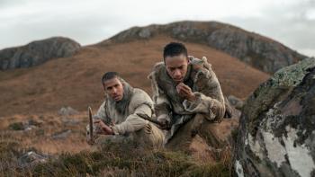 Out of Darkness Movie: Adem (Chuku Modu) and Geirr (Kit Young) crouch in the brush and rocks, both holding spears; Geirr examines something in his hand