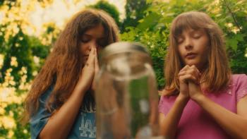 Over/Under: Two White girls standing outdoors, eyes closed, hands clasped in prayer, with green foliage behind