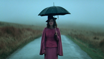 A woman in a pink period outfit stands alone on a misty country road in the rain, holding a black umbrella in Peaky Blinders: The Immortal Man