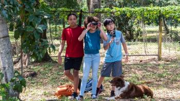 Pet Investigators: Three multiracial children with a dog laying beside them peer through binoculars and spy on people