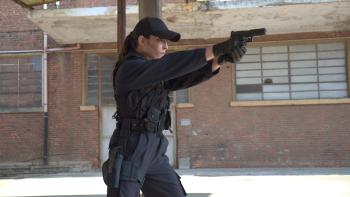 Assassin Club: A White woman dressed in black and wearing a cap aims her pistol