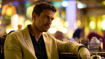 Fuze: A man in a cream blazer over a black shirt sits relaxed at what appears to be a bar or casino, with colorful bokeh lights in the background