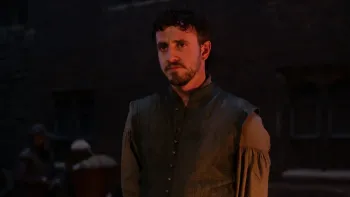 Hamnet: A man with dark curly hair and beard wearing period costume with a vest and billowing sleeves, lit dramatically against a dark background