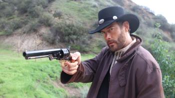 Place of Bones Movie: Villainous Bear John (Tom Hopper), in a black hat and brown duster, aims his gun