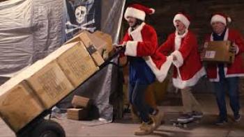Porch Pirates movie: Three adults dressed in Santa Claus robes and hats, center and center right, haul a dolly full of boxes in a warehouse