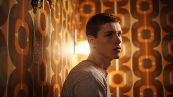 Postcards from London: Harris Dickinson stands in a dimly-lit room with orange patterned wallpaper, looking over his shoulder