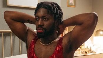 Pretty Red Dress: Natey Jones wears a red dress and lipstick as he reaches behind his head to fasten a gold necklace