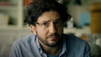 John Magaro looks concerned as he sits on a couch