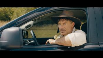 A man wearing an adventure hat sits in a black car with his arm propped outside the window. He is looking at something intently in the distance. 