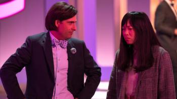 Quiz Lady movie: Jason Schwartzman in sports coat and bow tie on left stands next to and looks at Awkwafina in tweed sports coat on left